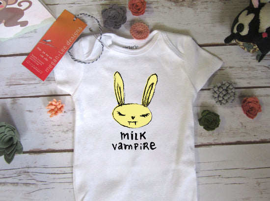 A white baby onesie with a yellow bunny graphic and the text "milk vampire" printed on it, along with a tag and some small decorative items on a wooden surface.