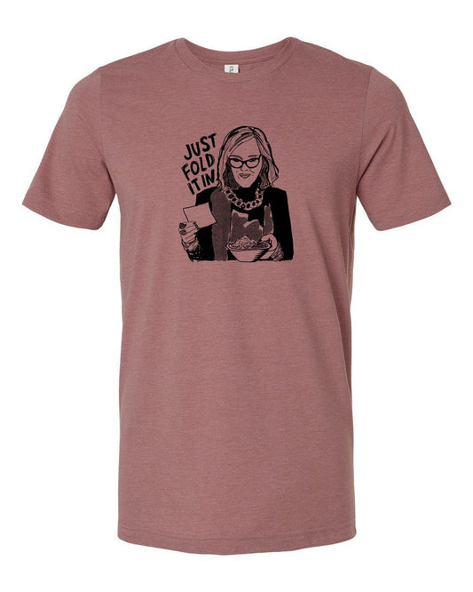 A graphic t-shirt design featuring a woman with glasses and a serious expression, holding a book and a cup of coffee. The text above the image says "JUST FOLD IT".