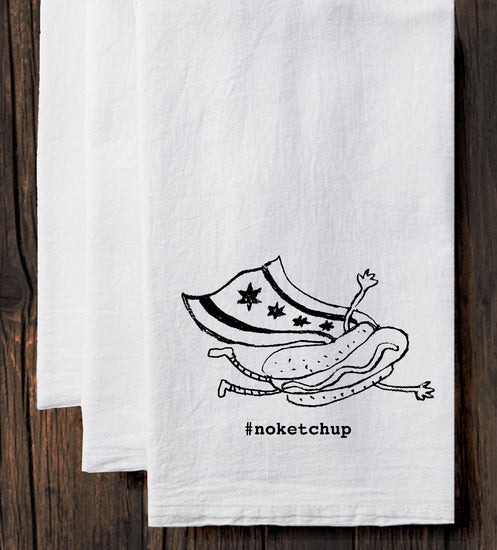 A white towel with a black and white illustration of a hot dog and a person wearing a superhero costume, along with the text "#noketchup".