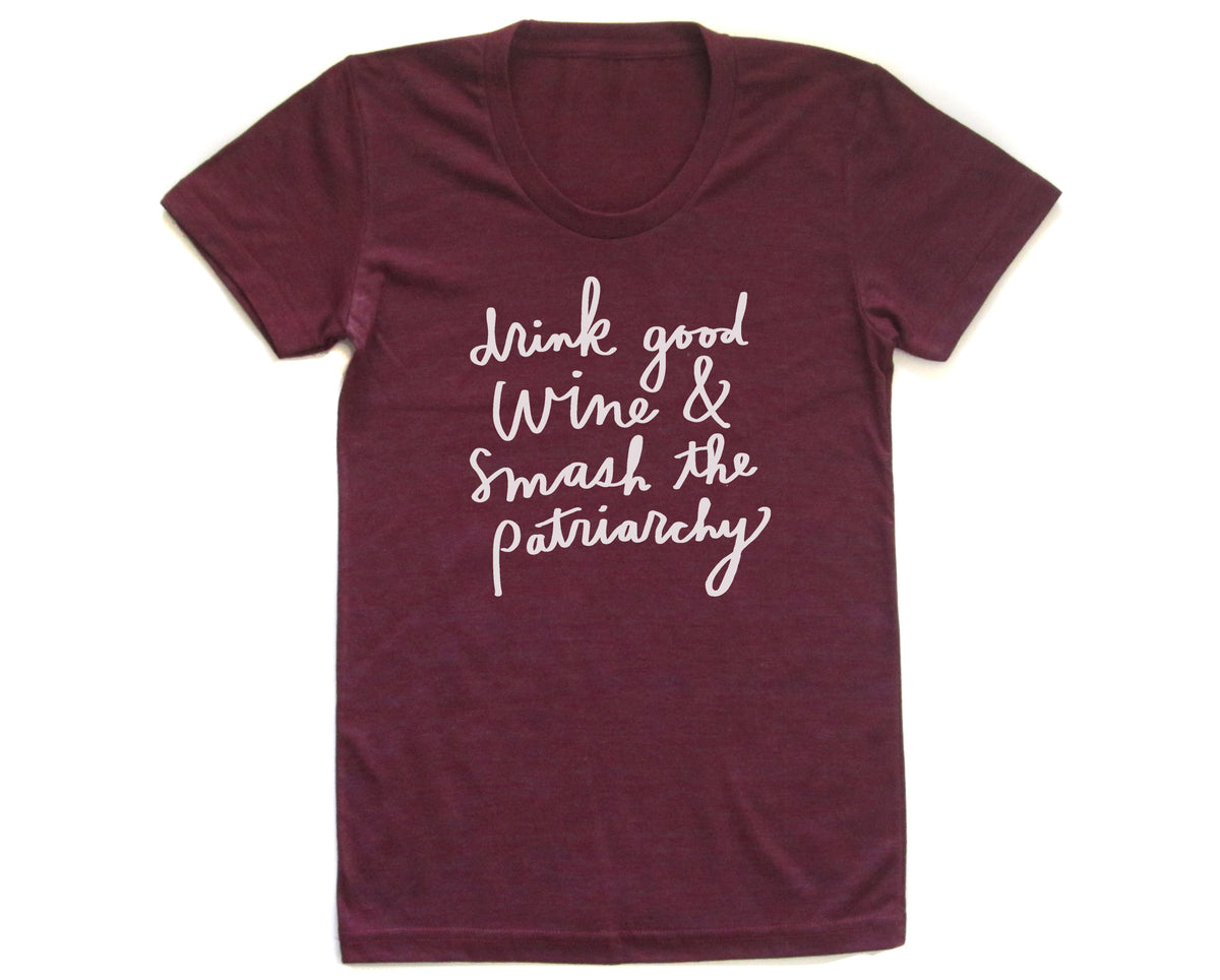 A burgundy-colored t-shirt with white text printed on it. The text reads "drink good wine & smash the patriarchy".