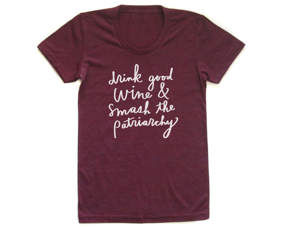 A burgundy-colored t-shirt with white text printed on it. The text reads "drink good wine & smash the patriarchy".