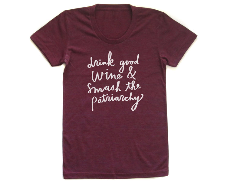 A burgundy-colored t-shirt with white text printed on it. The text reads "drink good wine & smash the patriarchy".