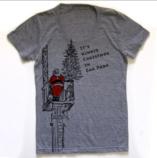 A gray t-shirt with a graphic design depicting a person in a red suit standing on a ladder and looking up at a Christmas tree. The text on the t-shirt reads "It's always Christmas in Oak Park".