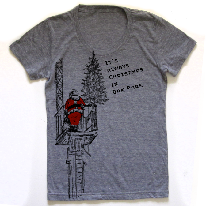 A gray t-shirt with a graphic design depicting a person in a red suit standing on a ladder and looking up at a Christmas tree. The text on the t-shirt reads "It's always Christmas in Oak Park".