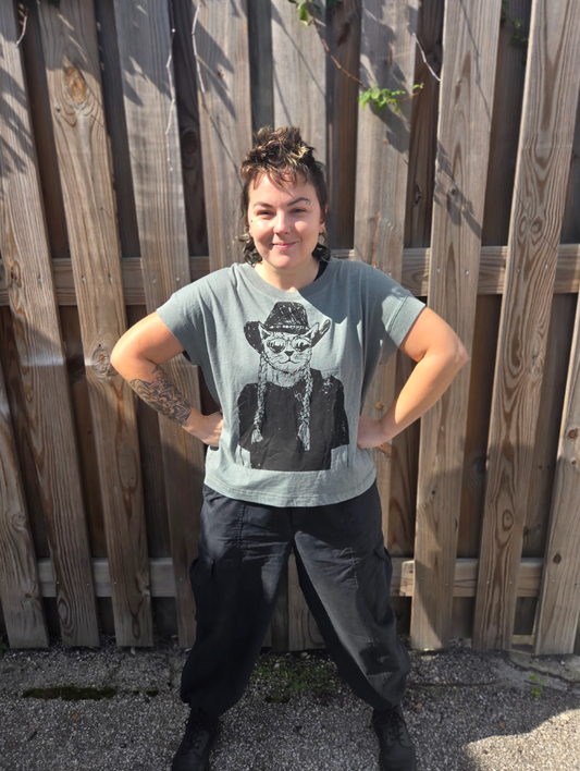 A person wearing a gray t-shirt with a graphic of a person wearing a cowboy hat, standing in front of a wooden fence.