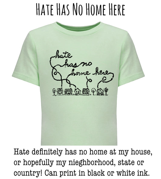 A light green t-shirt with a black handwritten message that reads " Hate has no home here" and a small illustration of houses below the text.