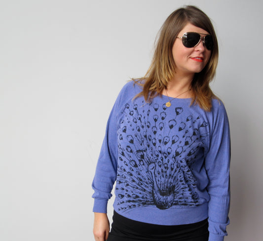 A woman wearing a purple sweater with a peacock design, sunglasses, and a necklace is posing for the camera.
