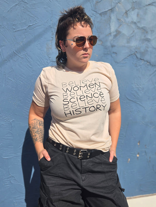 A person wearing a beige t-shirt with text printed on it, standing against a blue wall.