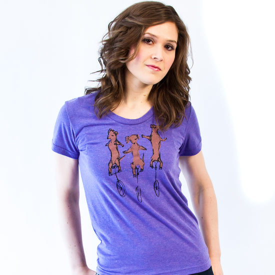 A woman wearing a purple t-shirt with a graphic of three dogs riding on unicycles.