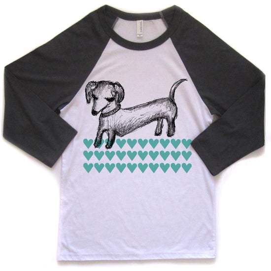 A white and black baseball-style t-shirt with a graphic design of a dachshund dog and a pattern of green hearts.