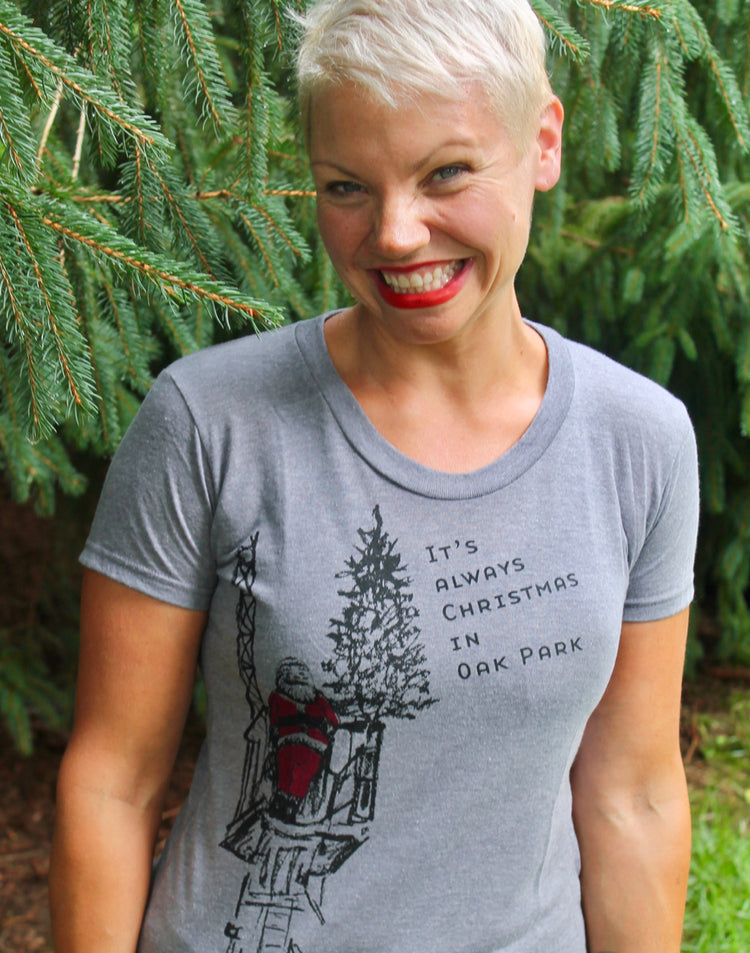 A smiling woman wearing a gray t-shirt with a Christmas tree graphic and text on it, standing in front of a pine tree.
