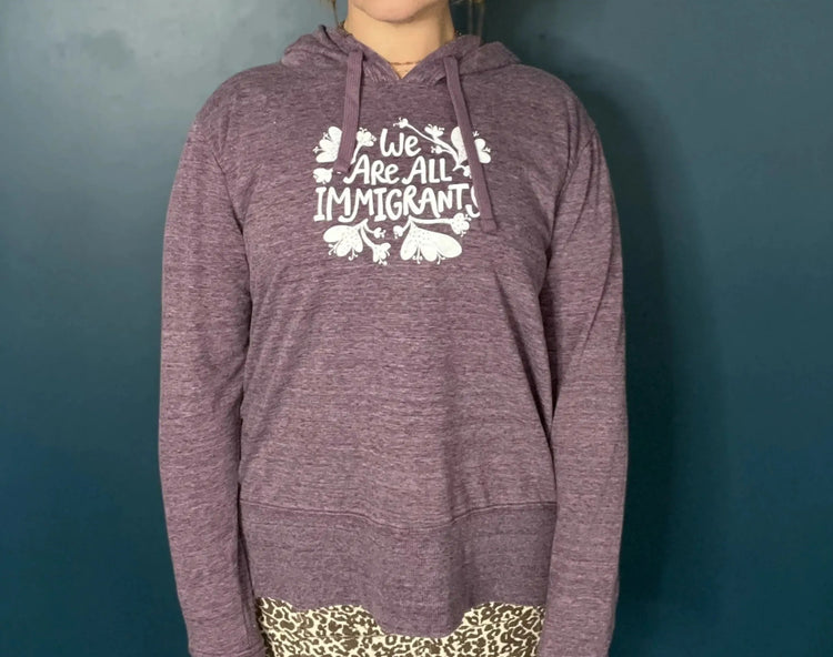 A woman wearing a purple hoodie with the text "We are all immigrants" stands against a blue background.