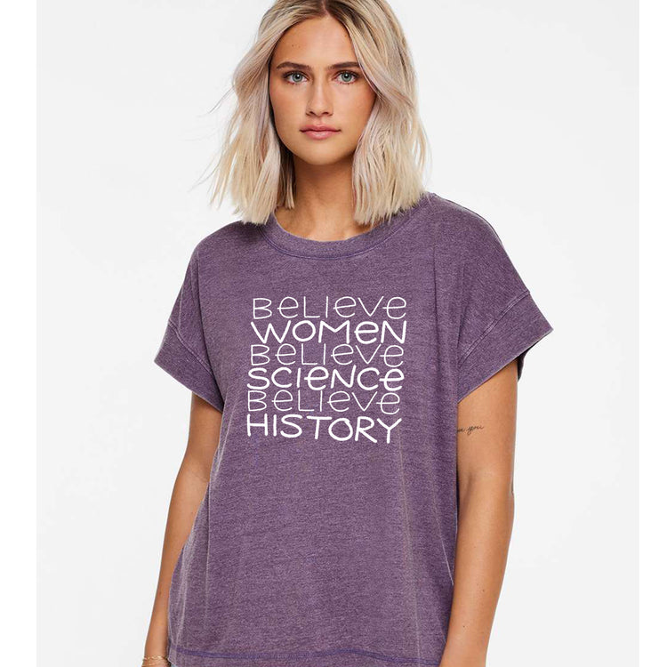 A woman with blonde hair wearing a purple t-shirt with the text "Believe Women Believe Science Believe History" printed on it.