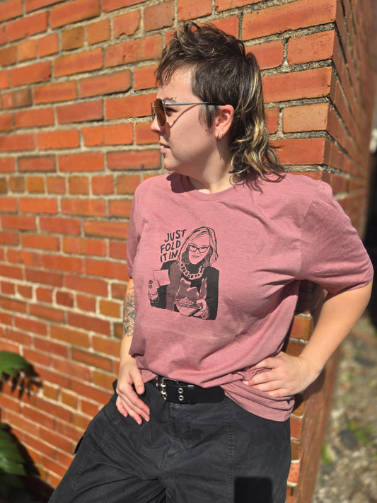 A person wearing a pink t-shirt with a graphic design is leaning against a brick wall.