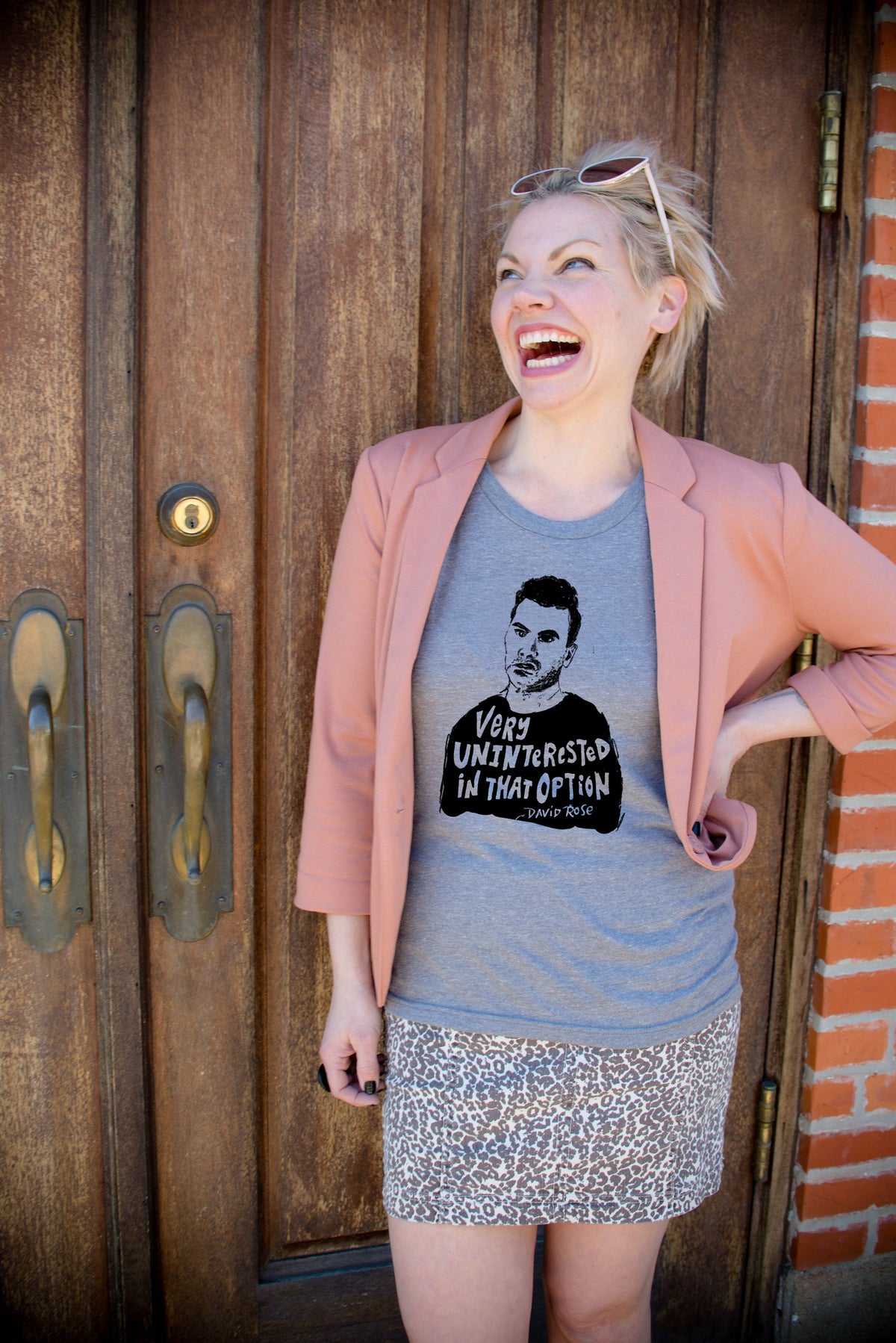 A woman wearing a pink blazer, a gray t-shirt with a graphic, and a leopard print skirt is standing in front of a wooden door.